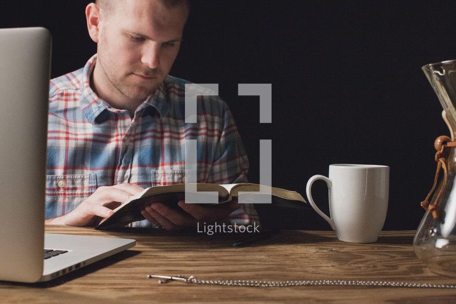 Man reading the Bible while sitting at a wooden table with a laptop computer and a coffee cup.