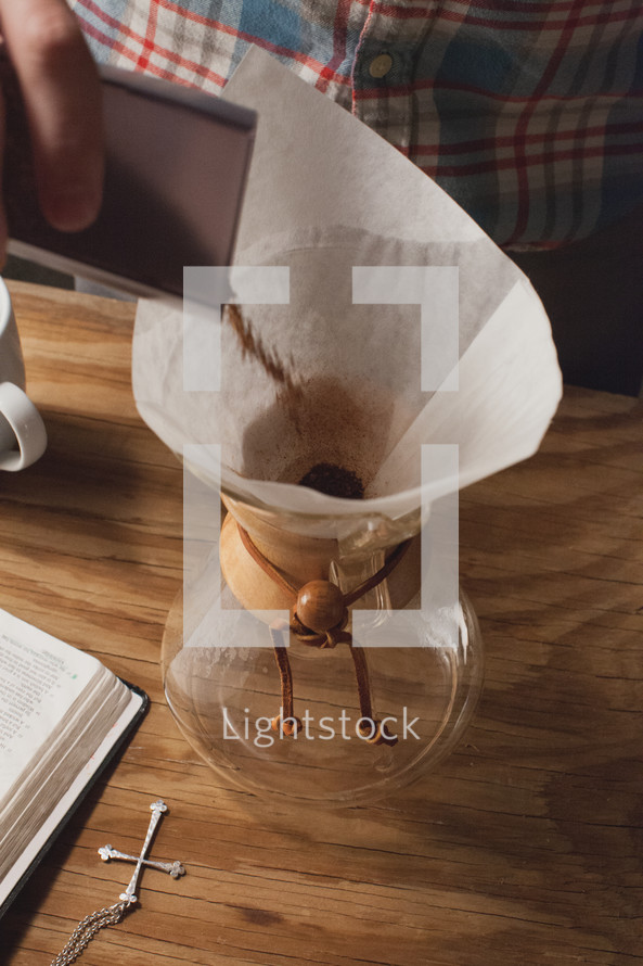 Coffee being poured into a chemex for brewing, next to a Bible and cross pendant.
