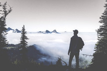 man standing on a mountain top in fog and clouds looking out at Mt Rainer