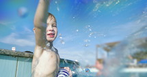Little boy in a swimming pool splashing water on the camera and laughing.