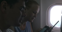 Man and a woman sitting on a plane using smart phones and tablets.