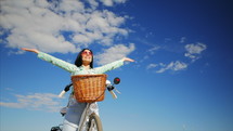 Happy young girl enjoying the sun sitting on a white vintage bicycle with basket. Hands up to the sky. Freedom concept. Carefree brunette girl. Slow motion.