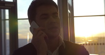 Young businessman talking on cell phone at the airport by a window at sunset.