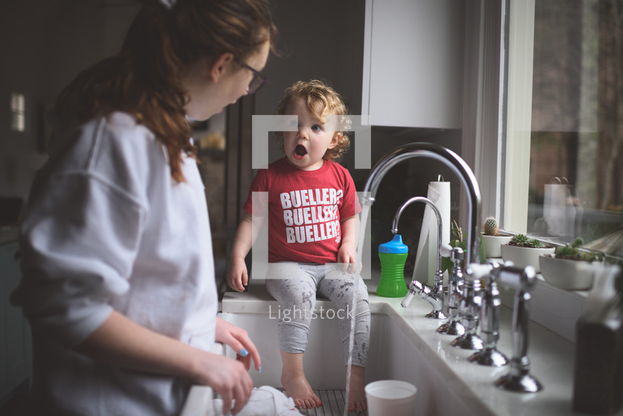 toddler playing in water at the kitchen sink while mom does dishes 