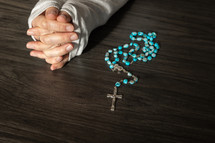 Woman's hands folded in prayer by a rosary on a dark wood table