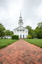Brick pathway through church yard leading to white church building with steeple surrounded by trees