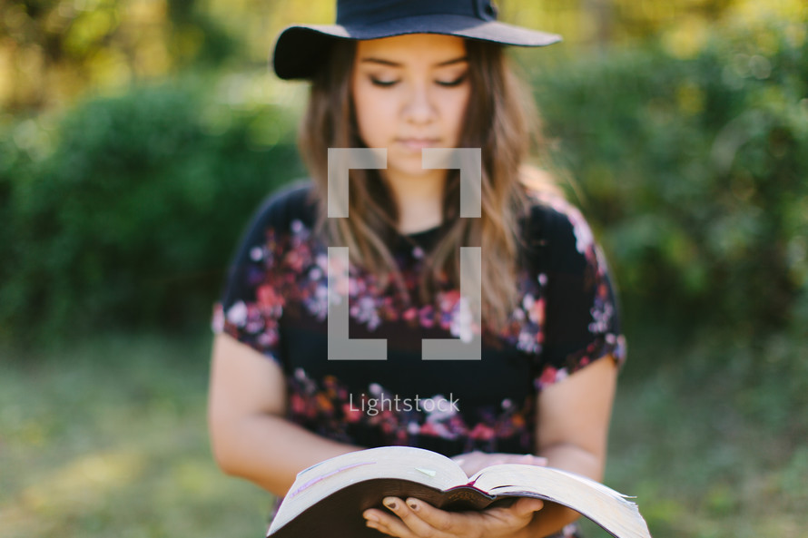 teen girl in a hat reading a Bible outdoors 