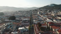 Long linear one-way street with many parked cars between the large residential blocks of the city of Oaxaca in Mexico with a high mountain range background. Low angle drone shot
