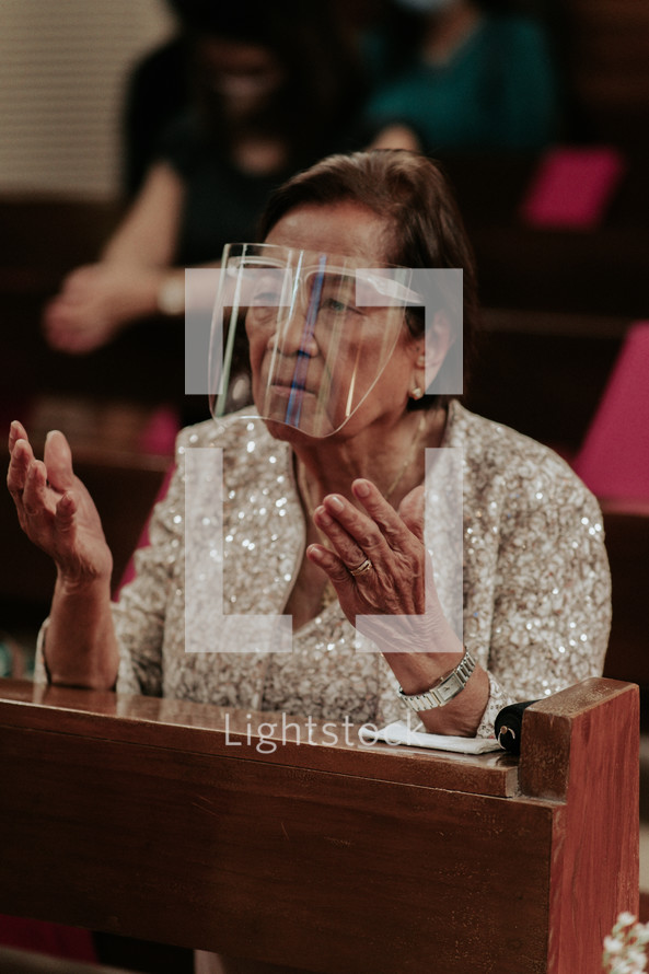 elderly woman in a face shield praying in church 