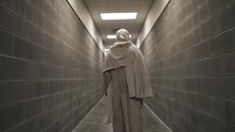 Jesus Christ dressed in white robes and tunic walking in dramatic, cinematic slow motion in the stone hallway of a prison or jail.