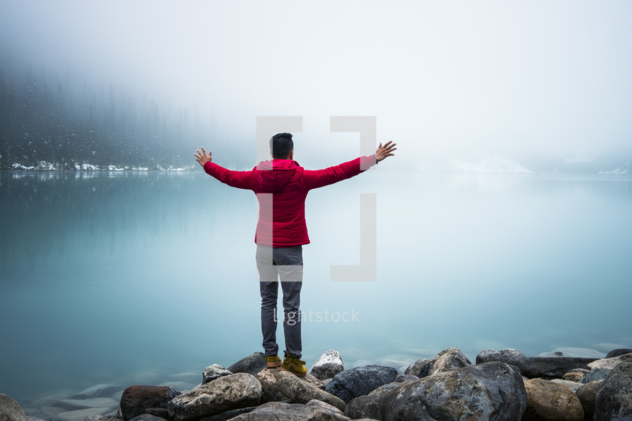 man standing on the shore of Lake Louise with hands raised 