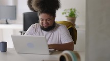 Black African American Man Having an Online Conversation on a Laptop Computer in Creative Office Environment. Happy Male is Browsing Social Media and Replying to Friends in Messenger.
