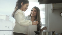 Little girl stirring boiling food in cooking pot and talking with joyful mother while preparing dinner together in domestic kitchen
