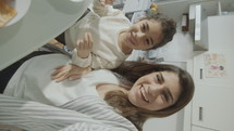 Joyful mother and little daughter having breakfast at kitchen table, waving on camera with a smile and chatting on web call in the morning at home. POV shot, vertical clip
