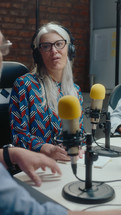 Senior woman in headphones talking into microphone, having conversation with co-hosts during audio podcast recording or radio broadcasting at studio. Vertical Clip