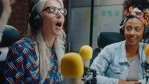 Senior and young female journalists in headphones sitting in recording studio, speaking into microphones and laughing, interviewing guest when co-hosting audio podcast