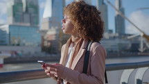 Young curly-haired black businesswoman standing on riverbank in the city, looking at downtown skyscrapers, texting on smartphone. Medium shot
