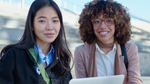 Young African-American and Asian businesswomen sitting outdoors with laptop, looking at camera and smiling happily. Medium shot, video portrait
