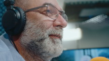 Senior male podcaster in headphones with gray beard talking into microphone in recording studio. Close-up shot, focus on foreground