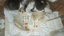 Mother helping daughter of preschool age to learn alphabet, both sitting at desk, using letters and word cards, girl writing in copybook. Top down shot, timelapse
