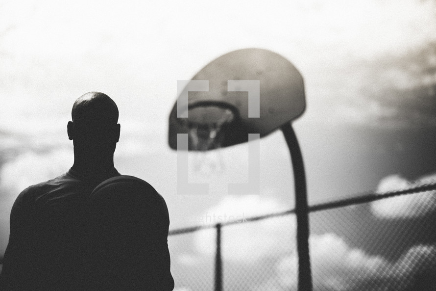 Silhouette of a man standing standing below a basketball hoop.