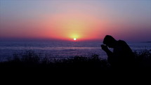 Silhouette of a man praying near the ocean water at sunset.