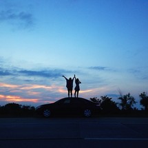 Two people stand on top of a car at dusk with arms raised to the sky.