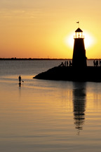 vertical shot of lighthouse at sunset with people at the base and a person on a paddleboard