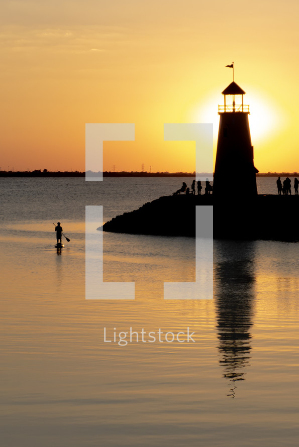 vertical shot of lighthouse at sunset with people at the base and a person on a paddleboard