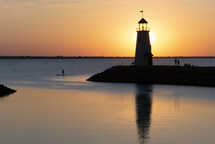 horizontal view of a silhouetted lighthouse at sunset with silhouettes of people watching the sunset by a peaceful lake