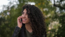 Beautiful young curly haired latin girl using her cell phone, outdoor. Portrait of relaxed young hispanic lady in green park, talking on phone