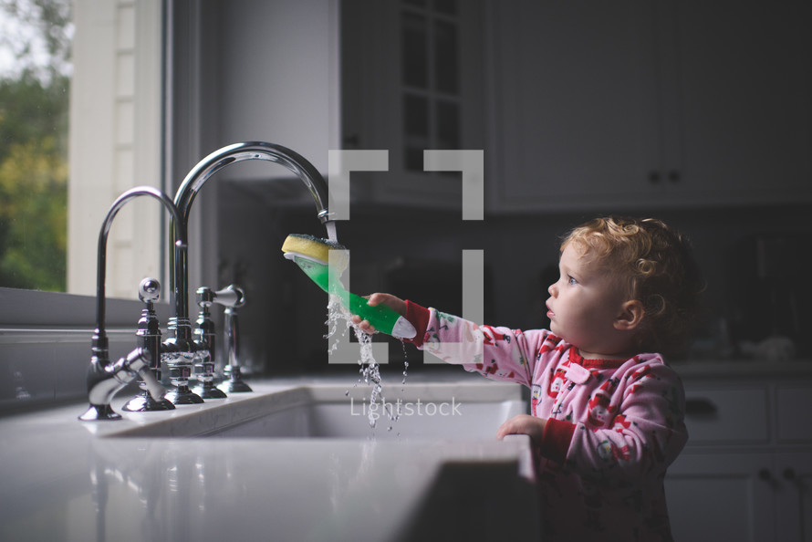 a toddler girl wetting a sponge in a kitchen sink 