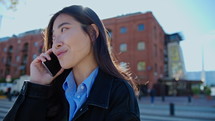 Young Asian woman standing outdoors on the street and talking on mobile phone during the day in the city. Handheld shot, lens flare
