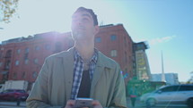 Young handsome man standing outdoors in the city, typing text message on smartphone and looking around at the street. Medium handheld shot
