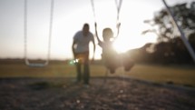 A father playing with his son at a park, pushing him on a swing in sunlight during sunset.