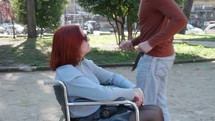 Woman in a wheelchair and man listening to music and dancing in a park.