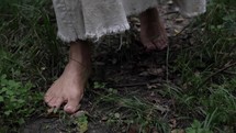 Slow motion tracking shot of the feet of Jesus Christ in white, tattered robe walking through grass in wooded area surrounded by trees.