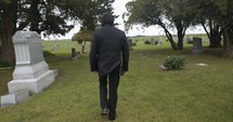 Sombre, sad young man in black suit walking in cinematic slow motion in cemetery through graveyard tombstones.