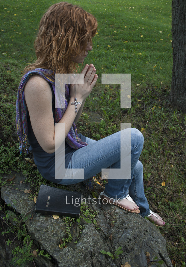 Teen girl holding cross, praying with Bible on fallen tree in park.