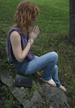 Teen girl holding cross, praying with Bible on fallen tree in park.