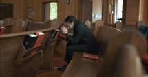 Man sitting next to bible in old church pew while praying, confessing, repenting of sin in cinematic slow motion.