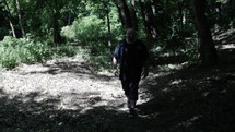 Middle aged man in black shirt with beard walking, hiking, meditating, praying contemplating in green, wooded area in trees with sunlight shining in cinematic, slow motion.