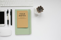 computer keyboard, field notes book, computer mouse, succulent plant, pens, and iPhone on a white background 