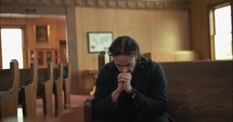 Young, emotional, anxious, and stressed man with long hair and black suit sitting in old church in worship and praying.