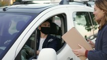 Delivery man wearing uniform, protection mask and gloves hand over a package to customer on the cargo van. Happy smiling female taking her parcel from the window of the car. Slow motion shot.
