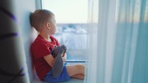 Child dreams concept. Friendship. Lonely little boy hugging teddy bear, looking out window, standing at home alone, happy child waiting for parents. Toddler child sitting at window sill, stay at home.