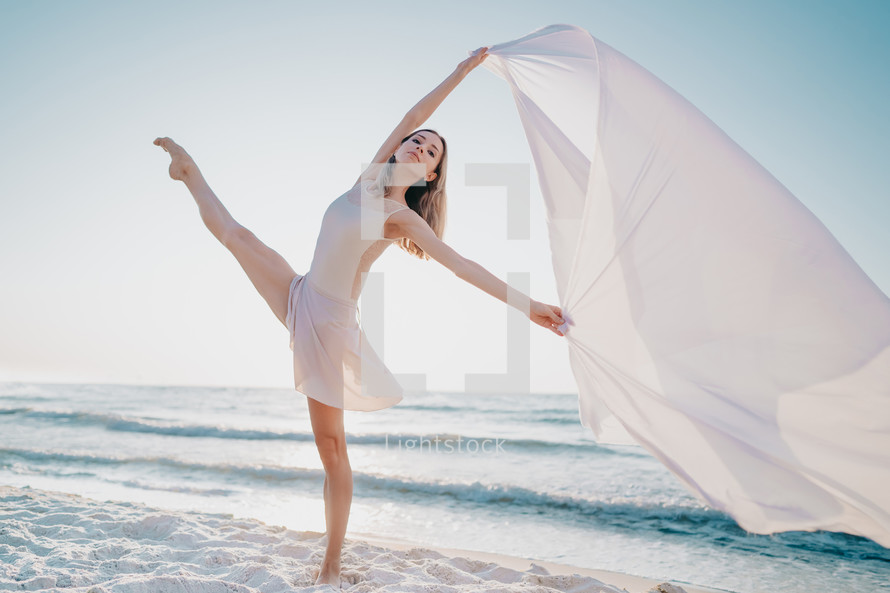 Young beautiful ballerina dancing ballet on seashore with huge silk fabric fluttering in wind.Concept of tenderness, lightness, art and talent in nature.

