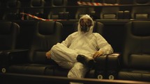 Relaxed man in white protection costume and respirator sitting alone at the cinema.