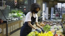 Mixed race grocery store employee in face mask and gloves putting fresh vegetables and fruits.