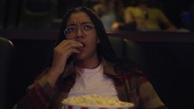 Diverse raced woman sitting in armchair watching a movie at the cinema, eating popcorn.